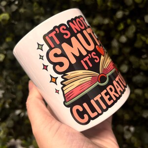 Op de afbeelding: Witte keramische mok met de tekst "It's not smut, it's cliterature" in een kleurrijk ontwerp. De mok heeft een illustratie van een open boek met een rood hart. Het ontwerp bevat sterren en een speels lettertype.