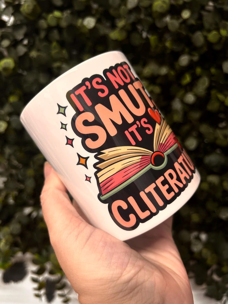 Op de afbeelding: Witte keramische mok met de tekst "It's not smut, it's cliterature" in een kleurrijk ontwerp. De mok heeft een illustratie van een open boek met een rood hart. Het ontwerp bevat sterren en een speels lettertype.