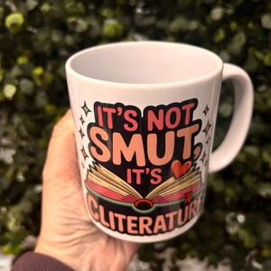 Op de afbeelding: Witte keramische mok met de tekst "IT'S NOT SMUT, IT'S CLITERATURE" in een kleurrijk ontwerp. Het ontwerp bevat een open boek met hart- en steraccenten. De mok heeft een gebogen handvat.