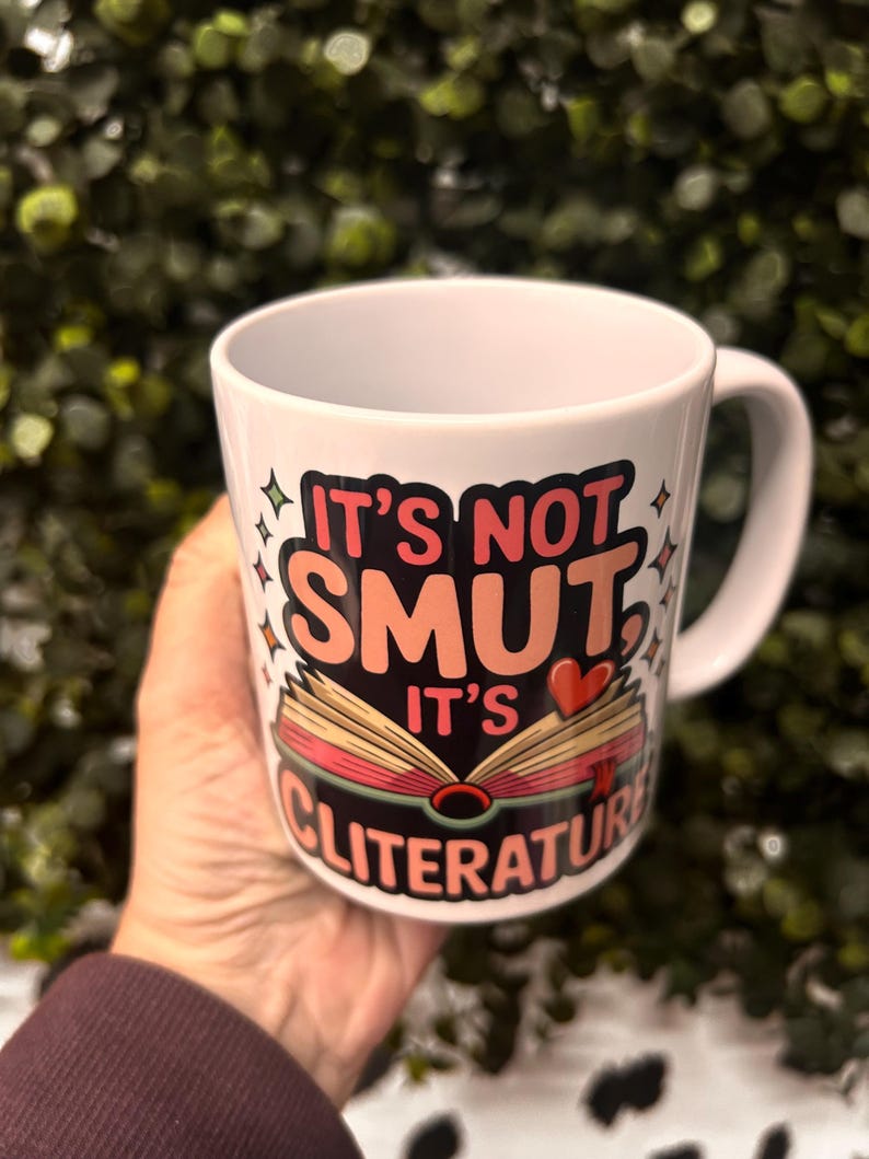 Op de afbeelding: Witte keramische mok met de tekst "IT'S NOT SMUT, IT'S CLITERATURE" in een kleurrijk ontwerp. Het ontwerp bevat een open boek met hart- en steraccenten. De mok heeft een gebogen handvat.