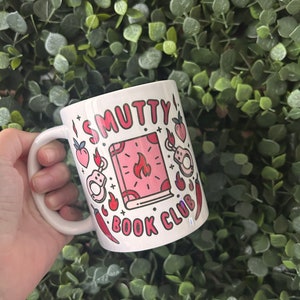 May include: White ceramic mug with a pink and red design. The mug features a book with flames, handcuffs, and chilli peppers. The text on the mug reads "SMUTTY BOOK CLUB".
