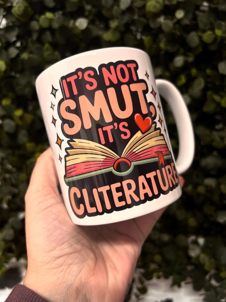Op de afbeelding: Witte keramische mok met de tekst "IT'S NOT SMUT IT'S CLITERATURE" in roze, perzik en rood. De mok heeft een illustratie van een open boek met een rood hart en een bladwijzer. De mok heeft een wit handvat.