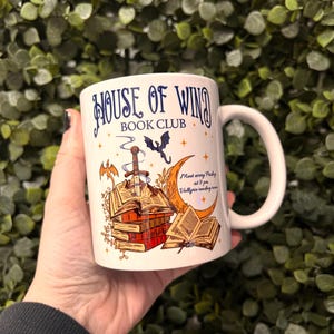 May include: White ceramic mug with a blue and gold design. The mug features a stack of books, a sword, a bat, and a crescent moon. The text "House of Wind Book Club" is printed on the mug. The text "Meet every Friday at 9 pm. Valkyrie reading room" is also printed on the mug.