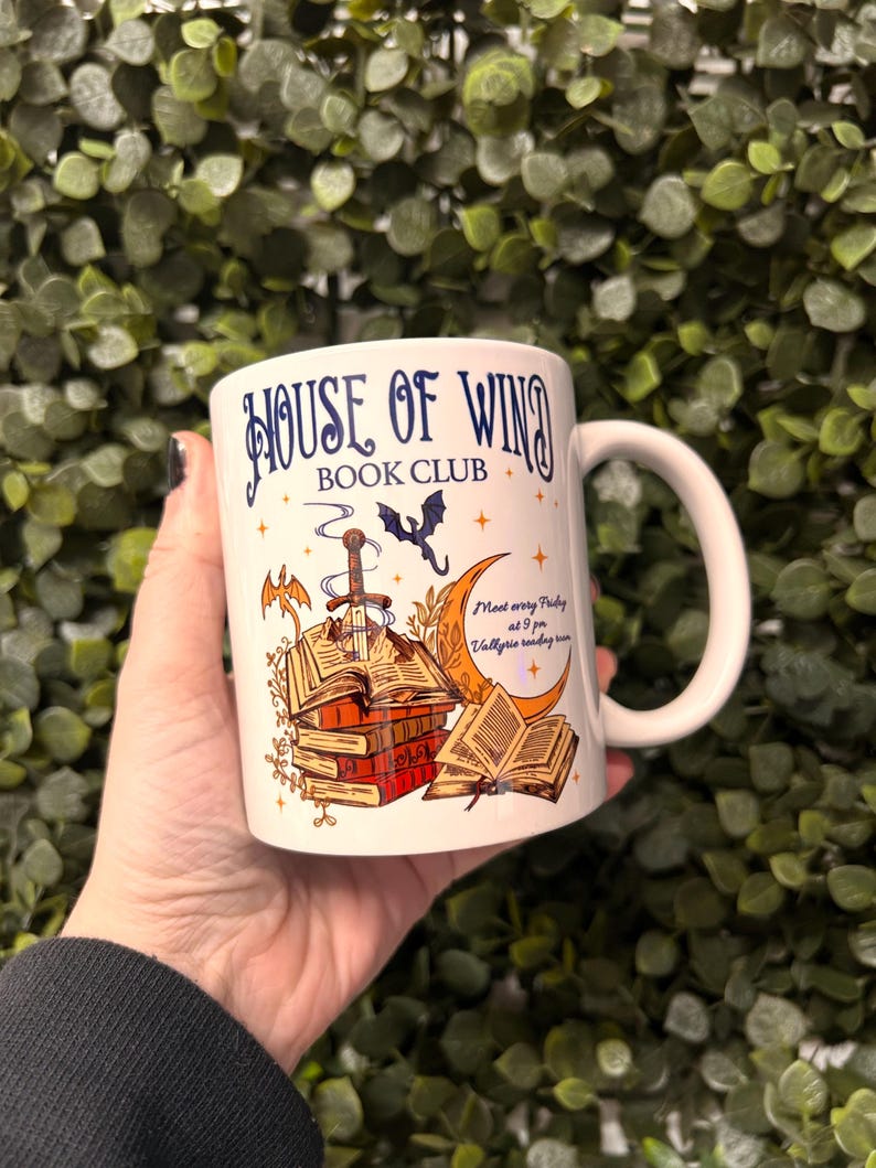 May include: White ceramic mug with a blue and gold design. The mug features a stack of books, a sword, a bat, and a crescent moon. The text "House of Wind Book Club" is printed on the mug. The text "Meet every Friday at 9 pm. Valkyrie reading room" is also printed on the mug.