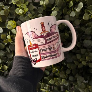 May include: White ceramic mug with a fantasy book theme. The mug features a red candle with the text "smells like... FANTASY", a book with the text "Chosen One", a book with the text "Fated Mates", and a book with the text "Mystical Creatures".