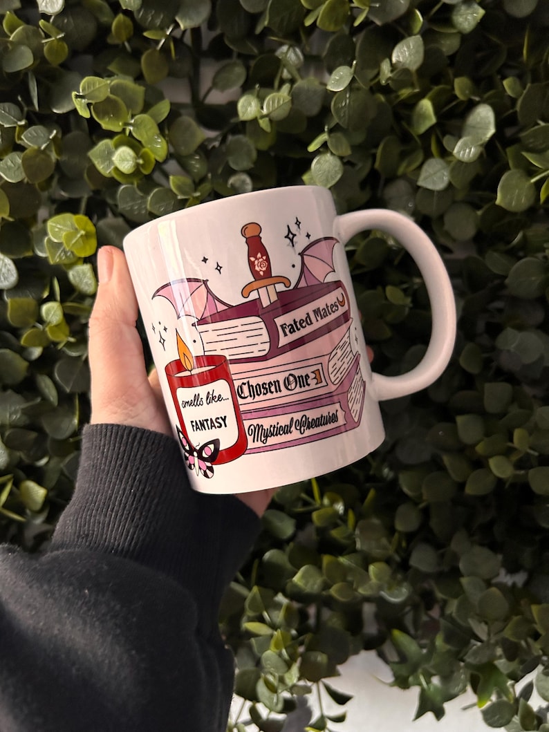May include: White ceramic mug with a fantasy book theme. The mug features a red candle with the text "smells like... FANTASY", a book with the text "Chosen One", a book with the text "Fated Mates", and a book with the text "Mystical Creatures".