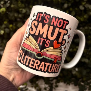 Op de afbeelding: Witte keramische mok met de tekst "IT'S NOT SMUT IT'S CLITERATURE" in roze, zwart en oranje. De mok heeft een illustratie van een open boek met een hart en sterren.