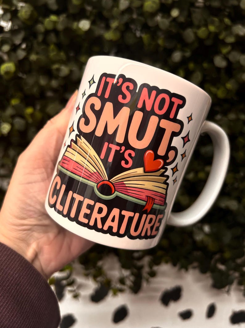 Op de afbeelding: Witte keramische mok met de tekst "IT'S NOT SMUT IT'S CLITERATURE" in roze, zwart en oranje. De mok heeft een illustratie van een open boek met een hart en sterren.