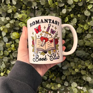 May include: White ceramic mug featuring a black and white illustration of a book, a sword, and a heart. The text "Romantasy Book Club" is printed on the mug.