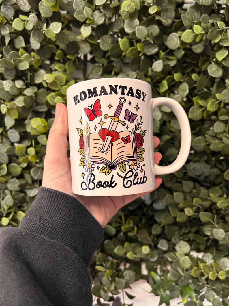 May include: White ceramic mug featuring a black and white illustration of a book, a sword, and a heart. The text "Romantasy Book Club" is printed on the mug.