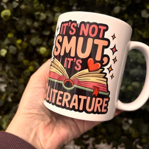 Op de afbeelding: Witte keramische mok met de tekst "IT'S NOT SMUT IT'S LITERATURE" in een kleurrijk ontwerp met een open boek, een rood hart en sterren. De mok heeft een gebogen handvat.
