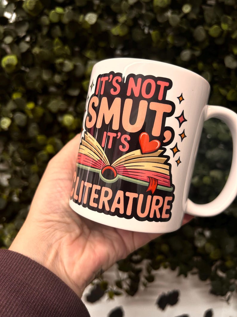 Op de afbeelding: Witte keramische mok met de tekst "IT'S NOT SMUT IT'S LITERATURE" in een kleurrijk ontwerp met een open boek, een rood hart en sterren. De mok heeft een gebogen handvat.