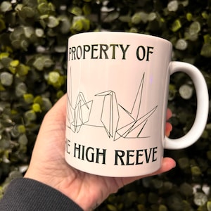 May include: White ceramic mug with the text "PROPERTY OF THE HIGH REEVE" in black. The mug features a geometric crane design. The mug has a white handle and is held in a hand.