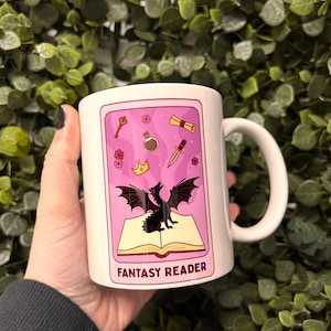 May include: White ceramic mug with a pink and black design. The design features a black dragon, a book, a crown, a key, a potion bottle, a dagger, and a scroll. The text "Fantasy Reader" is printed on the mug.