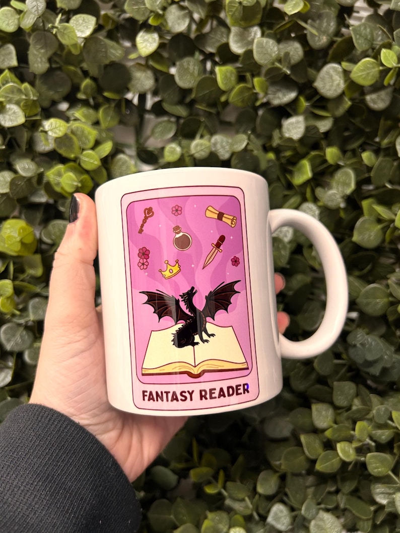 May include: White ceramic mug with a pink and black design. The design features a black dragon, a book, a crown, a key, a potion bottle, a dagger, and a scroll. The text "Fantasy Reader" is printed on the mug.