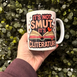 Op de afbeelding: Witte keramische mok met de tekst "IT'S NOT SMUT, IT'S CLITERATURE" in roze, zwart en rood. De mok heeft een afbeelding van een open boek met een hart. De mok heeft een wit oor en wordt vastgehouden voor een groene, bladerrijke achtergrond.