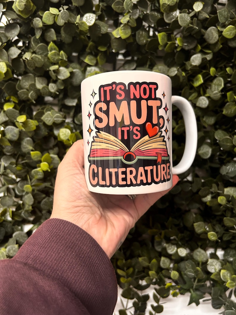 Op de afbeelding: Witte keramische mok met de tekst "IT'S NOT SMUT, IT'S CLITERATURE" in roze, zwart en rood. De mok heeft een afbeelding van een open boek met een hart. De mok heeft een wit oor en wordt vastgehouden voor een groene, bladerrijke achtergrond.