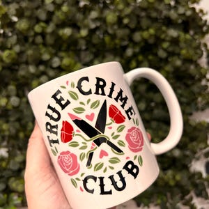 May include: White ceramic mug with a black design that says "True Crime Club" with a pair of crossed knives and roses in pink and red.