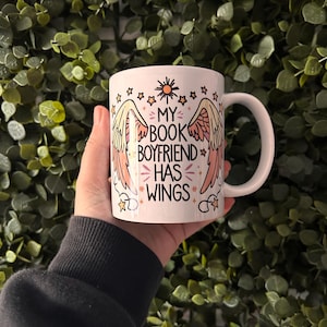 May include: White ceramic mug with a pink and yellow design featuring wings and the text "My Book Boyfriend Has Wings".