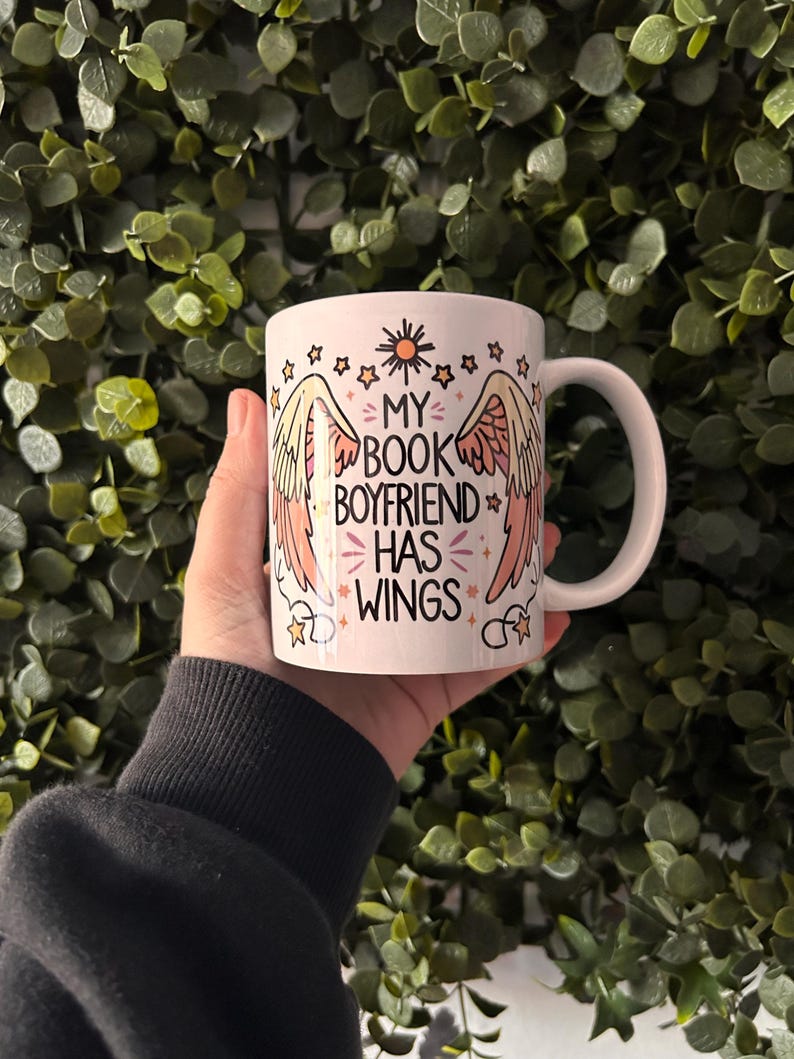 May include: White ceramic mug with a pink and yellow design featuring wings and the text "My Book Boyfriend Has Wings".