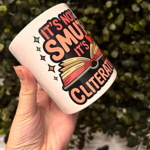 Op de afbeelding: Witte keramische mok met de tekst "It's not smut, it's cliterate" in een kleurrijk ontwerp. De mok heeft een boekafbeelding met rode, gele en roze pagina's. De mok wordt in een hand gehouden.