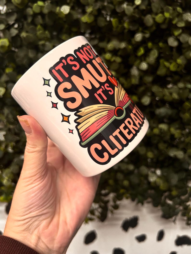 Op de afbeelding: Witte keramische mok met de tekst "It's not smut, it's cliterate" in een kleurrijk ontwerp. De mok heeft een boekafbeelding met rode, gele en roze pagina's. De mok wordt in een hand gehouden.