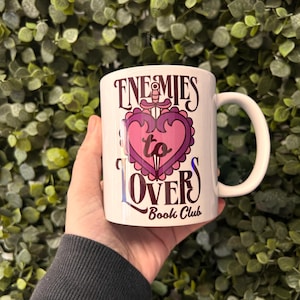 May include: White ceramic mug with a pink and purple heart design and the text "Enemies to Lovers Book Club".