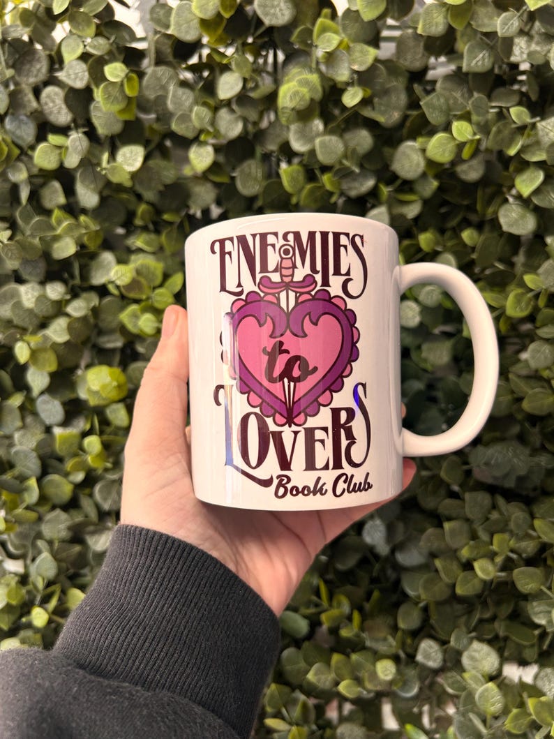 May include: White ceramic mug with a pink and purple heart design and the text "Enemies to Lovers Book Club".