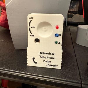 May include: A white Teleoicer Telephone Voice Changer with a volume dial, on/off switch, and colored buttons. The device has the words "Teleoicer Telephone Voice Changer" printed on the front.