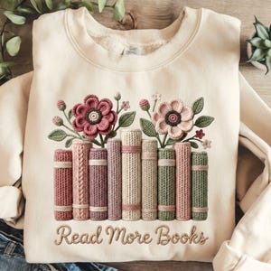 May include: A cream-coloured sweatshirt with an embroidered design. The design features a row of books and flowers. The books are in various colours, and the flowers are pink and green. The text "Read More Books" is embroidered below.