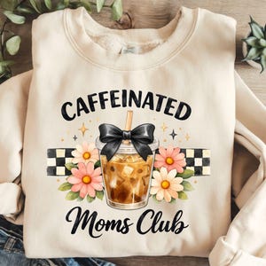 Könnte beinhalten: Ein cremefarbenes Sweatshirt mit Rundhalsausschnitt und einem Grafikdruck eines Eiskaffees in einem Plastikbecher mit schwarzer Schleife, umgeben von rosa und gelben Blumen und einem schwarz-weiß karierten Streifen, mit dem Text CAFFEINATED Moms Club.