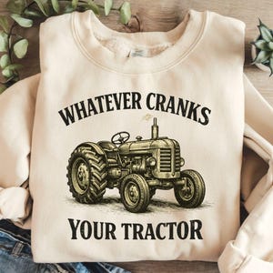 Könnte beinhalten: Cremefarbenes Sweatshirt mit einem Vintage-Traktor-Motiv. Der Text "WHATEVER CRANKS YOUR TRACTOR" ist oberhalb und unterhalb der Traktorillustration in Schwarz gedruckt. Der Traktor ist detailliert mit Rädern, Lenkrad und Schornstein.
