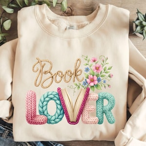 Op de afbeelding: Crèmekleurige sweatshirt met de woorden "Book Lover" in een grillig ontwerp. Het woord "Book" is in een touwachtige lettertype, terwijl "Lover" in een gebreide stijl is met een boek in het midden. Bloemenaccenten voegen kleur toe.