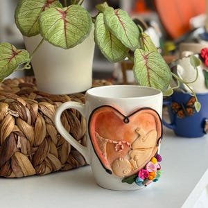 May include: A white ceramic mug featuring a three-dimensional clay sculpture of a fetus inside a heart-shaped womb. The sculpture includes a small umbilical cord and is decorated with a cluster of colorful miniature flowers at the bottom right.