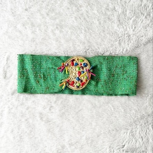 May include: A green headband with a gold-colored, oval-shaped embellishment. The embellishment is decorated with colorful beads and small tassels. The headband is made of a textured fabric and is suitable for various occasions.