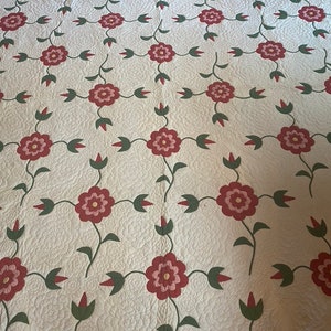Op de afbeelding: Een wit quilt met een herhalend patroon van rode bloemen met roze centra en groene bladeren. Het quilt is gestikt met een klein ruitpatroon.