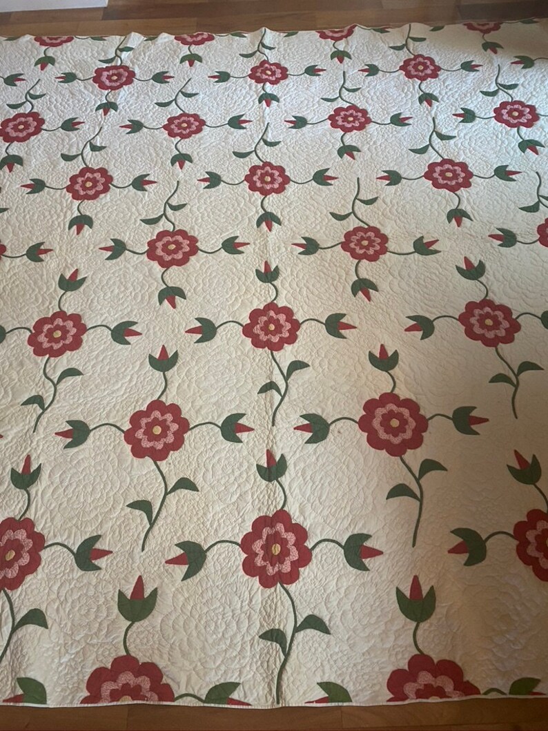 Op de afbeelding: Een wit quilt met een herhalend patroon van rode bloemen met roze centra en groene bladeren. Het quilt is gestikt met een klein ruitpatroon.