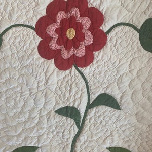 Op de afbeelding: Een vintage quilt met een grote rode bloem en groene bladeren. De quilt is gestikt in een patroon van kleine cirkels.