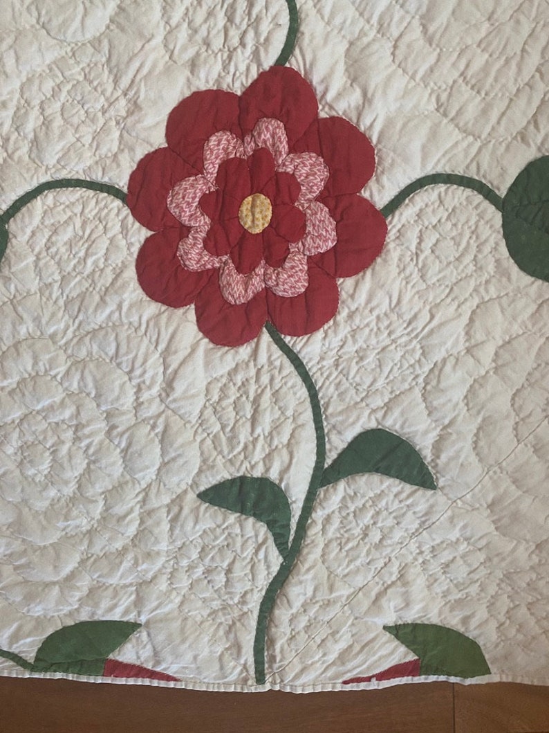 Op de afbeelding: Een vintage quilt met een grote rode bloem en groene bladeren. De quilt is gestikt in een patroon van kleine cirkels.