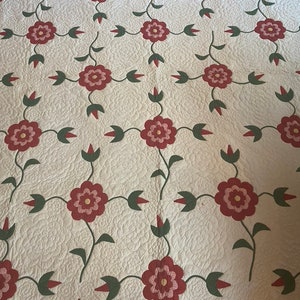 Op de afbeelding: Een wit quilt met een herhalend patroon van rode bloemen met roze centra en groene bladeren. Het quilt is gestikt met een klein ruitpatroon.