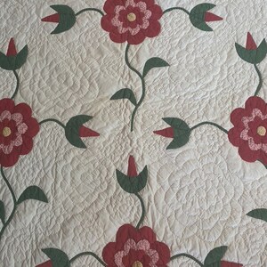 Op de afbeelding: Een wit quilt met een herhalend patroon van rode en groene bloemen. De bloemen zijn gemaakt van stof en zijn gequilt in een eenvoudig ontwerp.