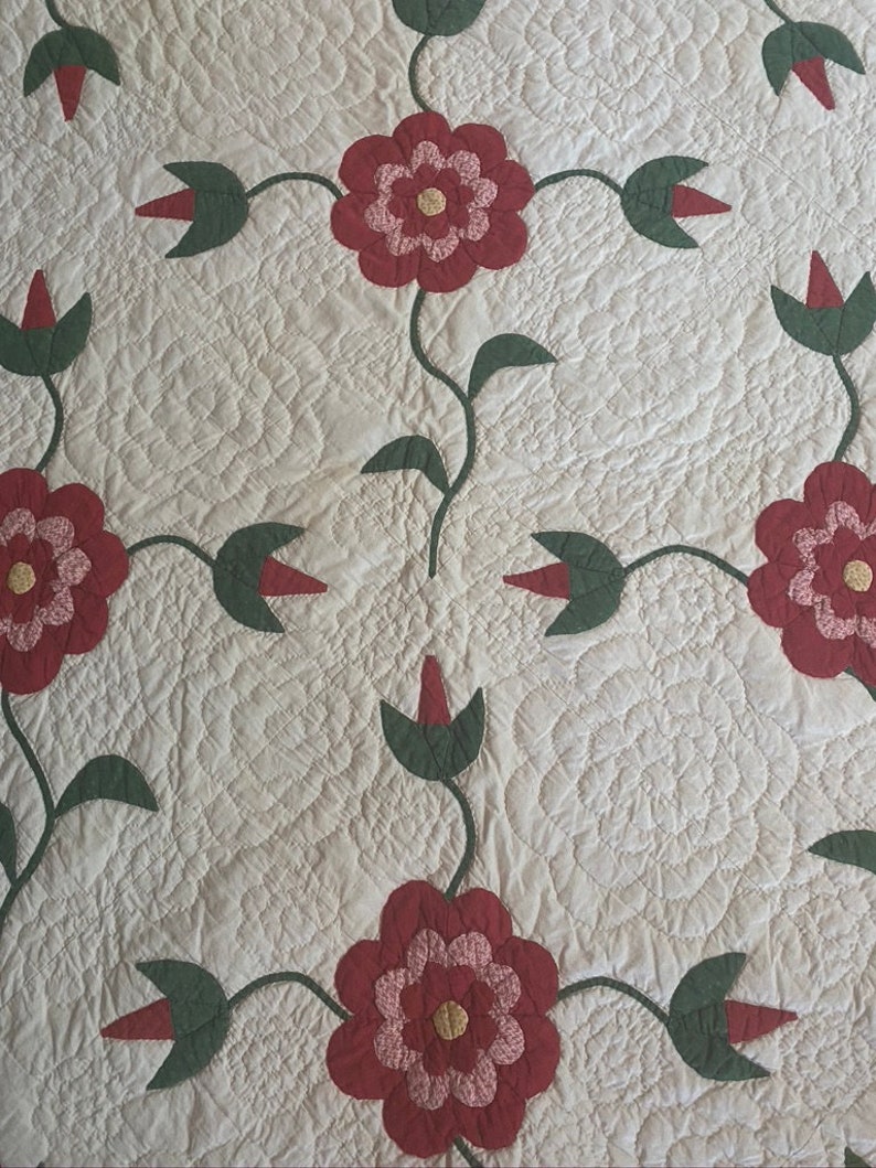 Op de afbeelding: Een wit quilt met een herhalend patroon van rode en groene bloemen. De bloemen zijn gemaakt van stof en zijn gequilt in een eenvoudig ontwerp.