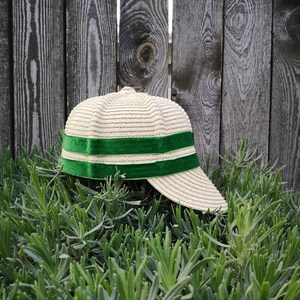 May include: A straw-colored baseball cap with a green velvet band. The cap is set against a weathered wooden fence and surrounded by green plants. The cap's brim is curved, and the green band adds a pop of color.