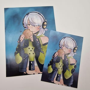 May include: Two art prints featuring an anime-style character with white hair and headphones, eating a burger. The character wears a black and green outfit. The background is a gradient of blue. The prints are on a white surface.