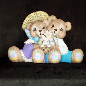 May include: Two ceramic teddy bears holding a basket of Easter eggs. The bears are wearing blue and purple outfits and have painted details. The basket is filled with colorful eggs and a white bunny.