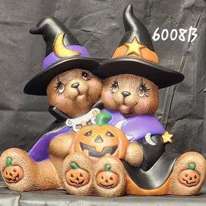 May include: A ceramic figurine of two brown bears dressed as witches, wearing black hats with a yellow moon and star. They hold a central orange jack-o'-lantern, with smaller pumpkins on their feet. The number 6008B is printed in the upper right.