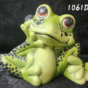 Puede incluir: Figura de rana de cerámica verde con manchas negras, apoyando la barbilla en la mano. La rana tiene ojos grandes con detalles en naranja y negro. El número "1061D" es visible en la esquina superior derecha.