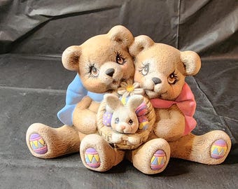 Könnte beinhalten: Zwei braune Teddybären halten einen Korb mit einem weißen Hasen darin. Die Bären tragen blaue und rosa Schals mit Osterei-Designs. Der Hase hält eine rosa und weiße Blume.