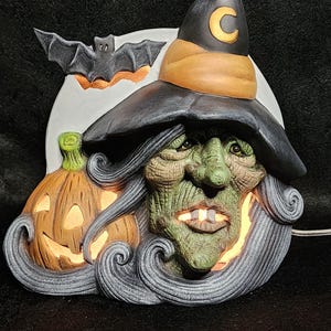 May include: A ceramic Halloween witch figurine with a black hat, green face, and long gray hair. The witch is holding a jack-o'-lantern and a bat is flying above her head.