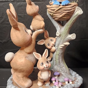 Puede incluir: Una figurita de cerámica de una familia de conejos con un nido de pájaro en un tronco de árbol. Los conejos son marrones y blancos, y los pájaros son azules. La base de la figurita está decorada con pequeñas setas.